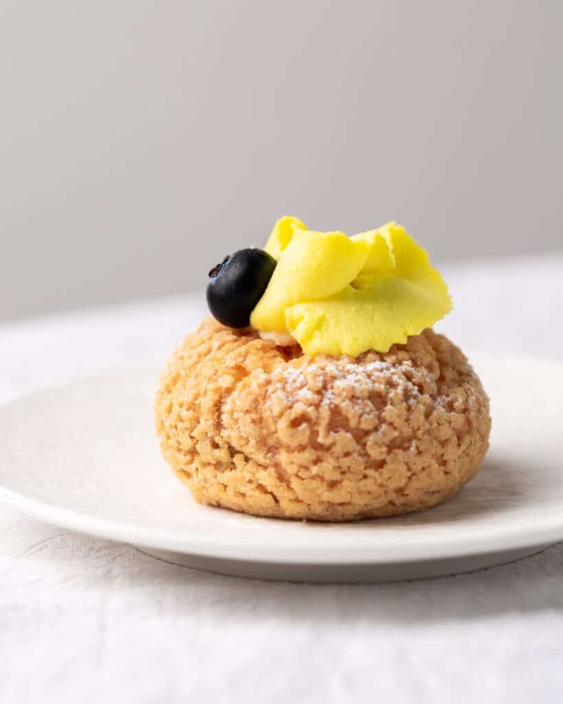 cream puff on a white plate topped with a blueberry and yellow cream