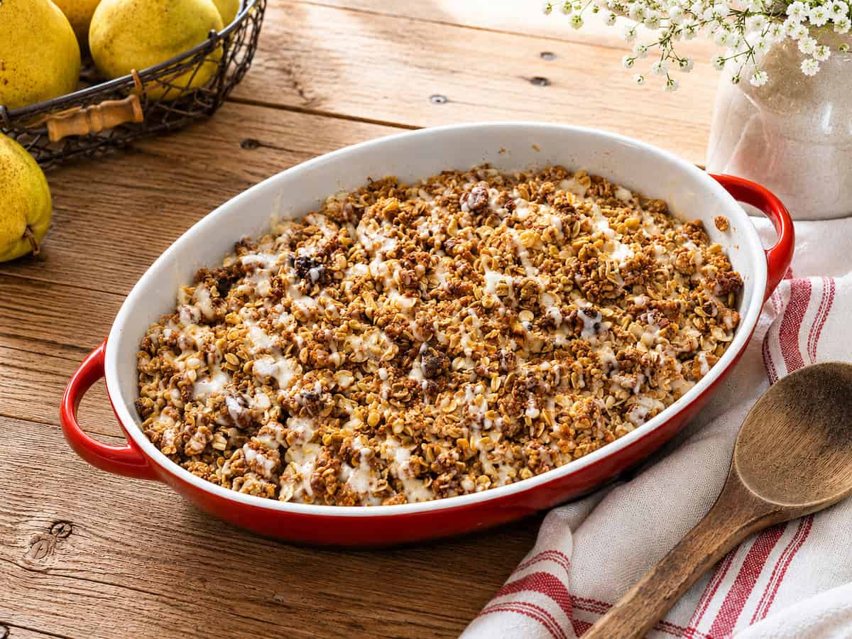 Brown Butter Honey Pear Crisp on a wooden table with a white towel next to it with a wooden spoon sitting on it. There's a black basket with a pear in it to the upper left.