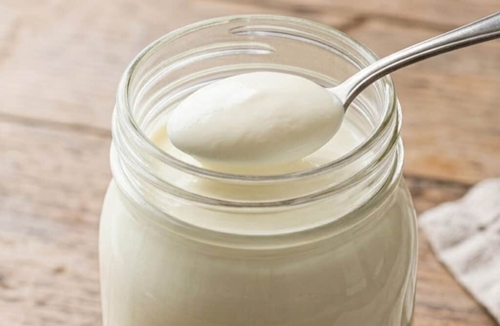 jar of sour cream on a wooden table with a metal spoon scooping some out.