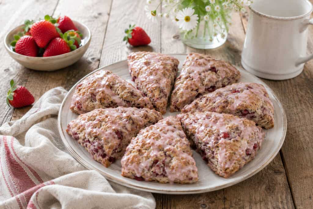 a white plant with 6 strawberry buttermilk scones drizzled with strawberry glaze on a wooden table with a small white bowl of strawberries beside it and a white cup and clear vase of daisies behind it.