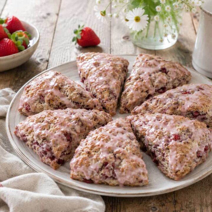 a white plant with 6 strawberry buttermilk scones drizzled with strawberry glaze on a wooden table with a small white bowl of strawberries beside it and a white cup and clear vase of daisies behind it.
