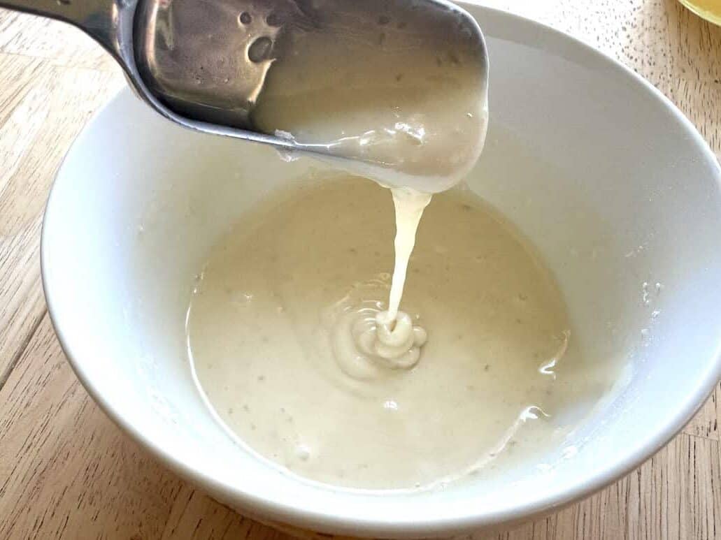 stainless steel tablespoon drizzling vanilla honey glaze into a white bowl that sits on a wooden table