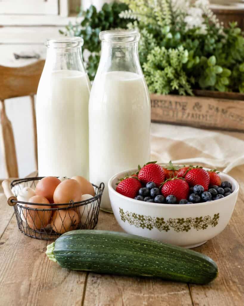 two milk bottles, a white bowl with strawberries and blueberries, a small black basket of brown and white eggs, and a zucchini sitting on a wooden table with a short wooden crate of plants in the background