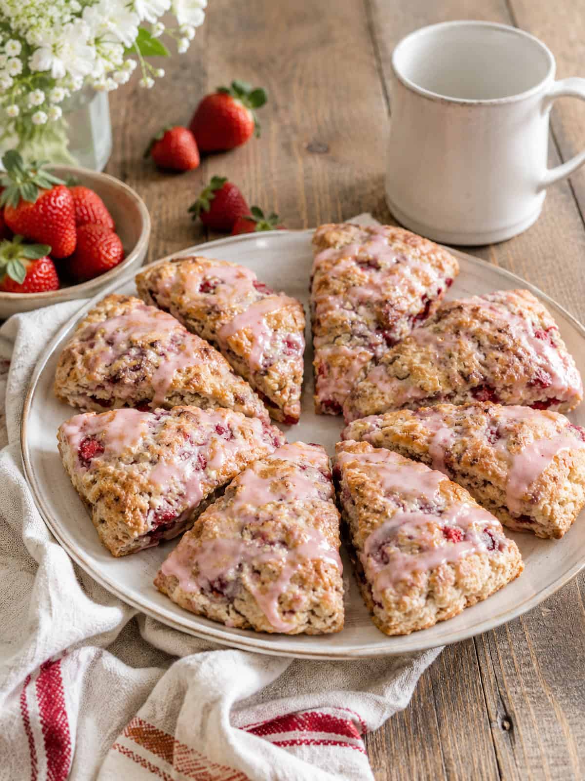a white plant with 6 strawberry buttermilk scones drizzled with strawberry glaze on a wooden table with a some strawberries and a white mug behind it.