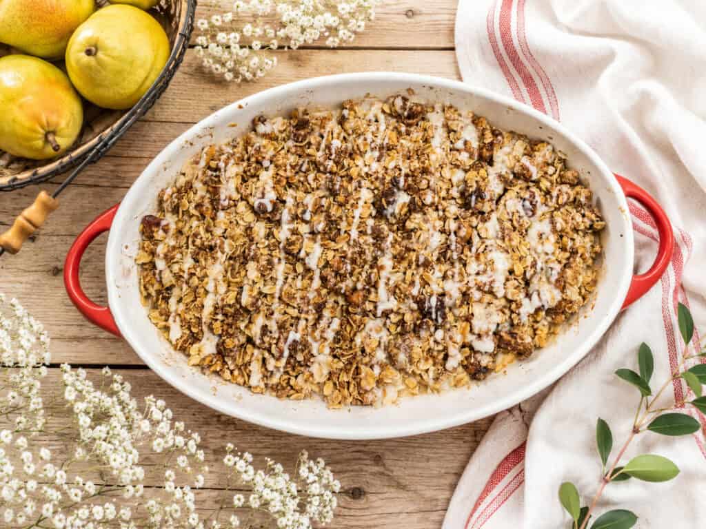 Brown Butter Honey Pear Crisp on a wooden table with a red and white towel next to it. There's a black basket with a pear in it to the upper left, some small white flowers to the lower left and upper middle, and some leaves to the lower right.
