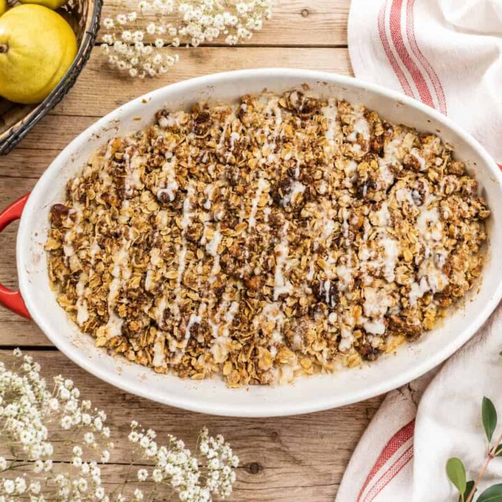 brown butter honey pear crisp in a white and red baking dish. It's sitting on a light wooden table with a white and red towel to the right, dried white flowers above it and a pear in a black basket to the upper left.