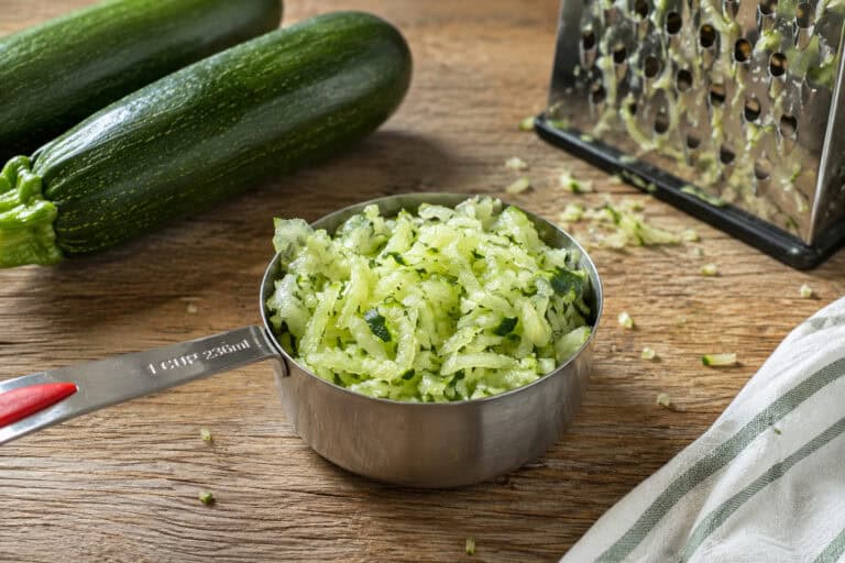 stainless steel one cup measuring cup of shredded zucchini on a wooden table. 2 zucchini are to the top left of the cup, a box grater with crumbs of zucchini on it is to the upper right, and a white and gray kitchen towel is to the bottom right. Bits of shredded zucchini are on the able around the measuring cup.