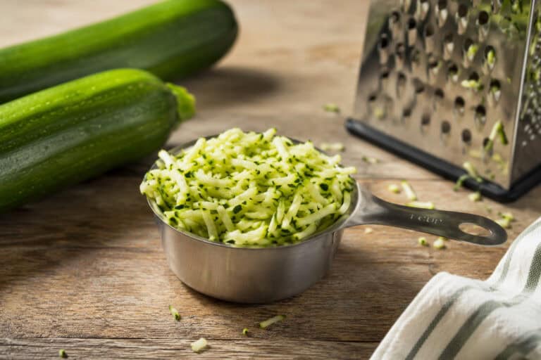 stainless steel one cup measuring cup of shredded zucchini on a wooden table. 2 zucchini are to the top left of the cup, a box grater with crumbs of zucchini on it is to the upper right, and a white and gray kitchen towel is to the bottom right. Bits of shredded zucchini are on the able around the measuring cup.