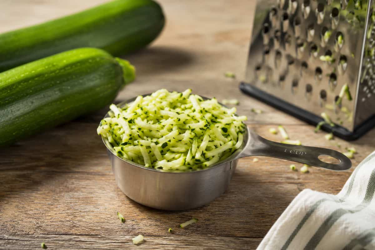 stainless steel one cup measuring cup of shredded zucchini on a wooden table. 2 zucchini are to the top left of the cup, a box grater with crumbs of zucchini on it is to the upper right, and a white and gray kitchen towel is to the bottom right. Bits of shredded zucchini are on the able around the measuring cup.