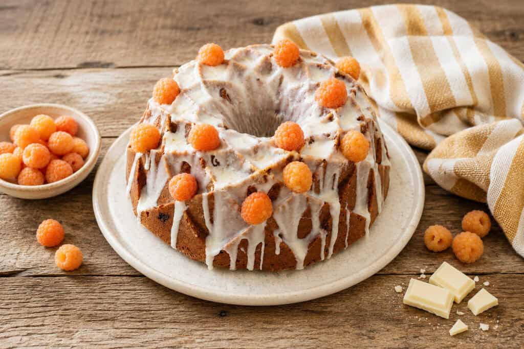Golden Raspberry white chocolate bundt cake topped with white chocolate glaze and fresh golden raspberries on a white plate sitting on a wooden table. A gold and white striped towel is to the right, golden raspberries in a white bowl are to the left. A few berries and a couple of pieces of white chocolate surround the bottom of the plate.