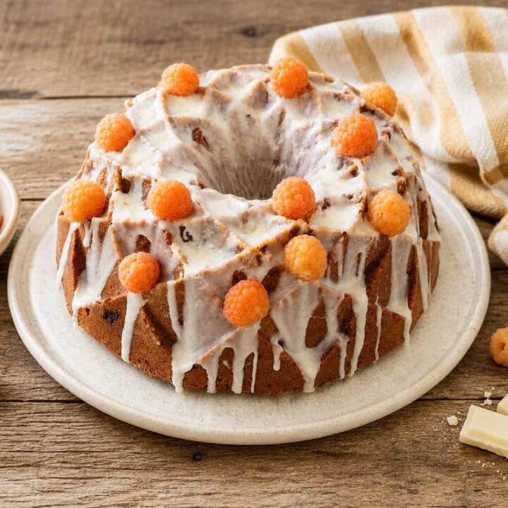 Golden Raspberry white chocolate bundt cake topped with white chocolate glaze and fresh golden raspberries on a white plate sitting on a wooden table. A gold and white striped towel is to the right, golden raspberries in a white bowl are to the left. A few berries and a couple of pieces of white chocolate surround the bottom of the plate.