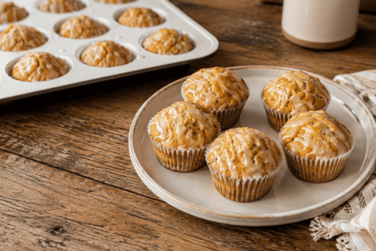 5 orange zucchini muffins on a white plate sitting on a wooden table with a tan and white kitchen towel to the right. A white muffin pan filled with more muffins is to the upper left of the plate.