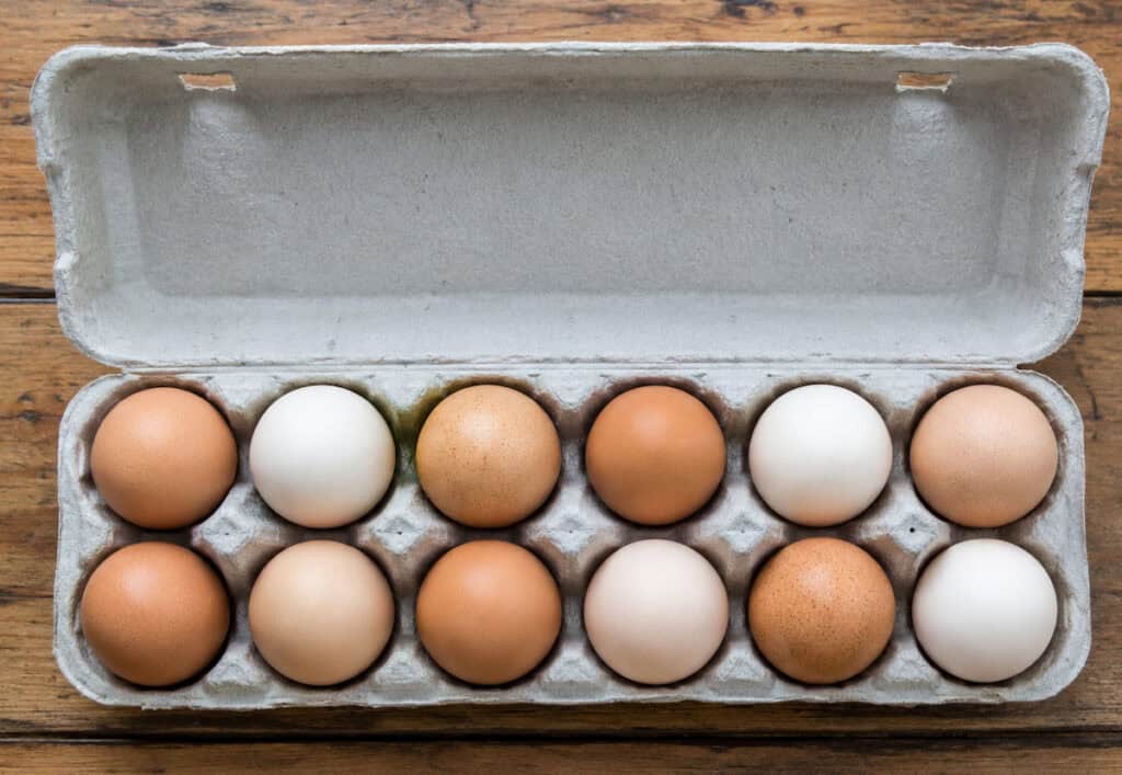 1 dozen eggs in white and varying shades of brown in a gray cardboard egg carton on a wooden table.