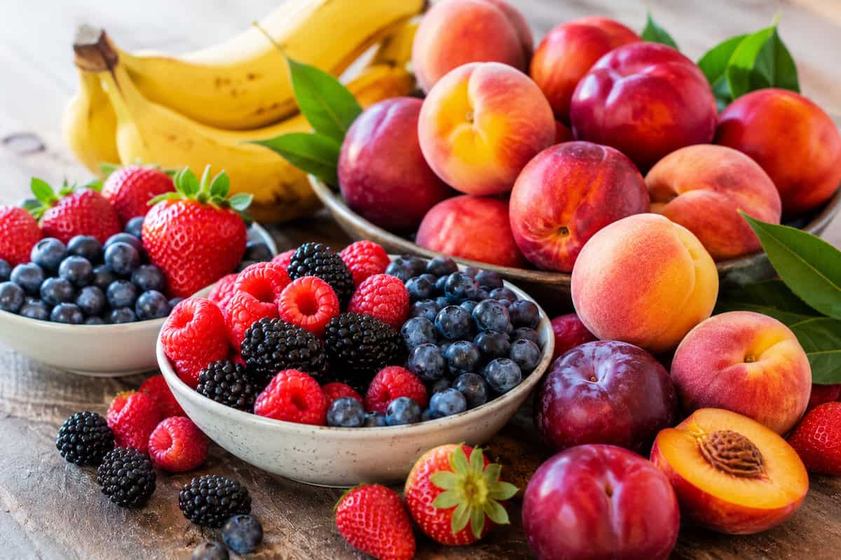 a white bowl of strawberries and blueberries, a white bowl of blueberries, raspberries, and blackberries, and a bowl of peaches and plums on a wooden table. A bunch of bananas are behind the bowls and more peaches, plumbs, strawberries, blackberries, and raspberries surround the bowls on the right side.