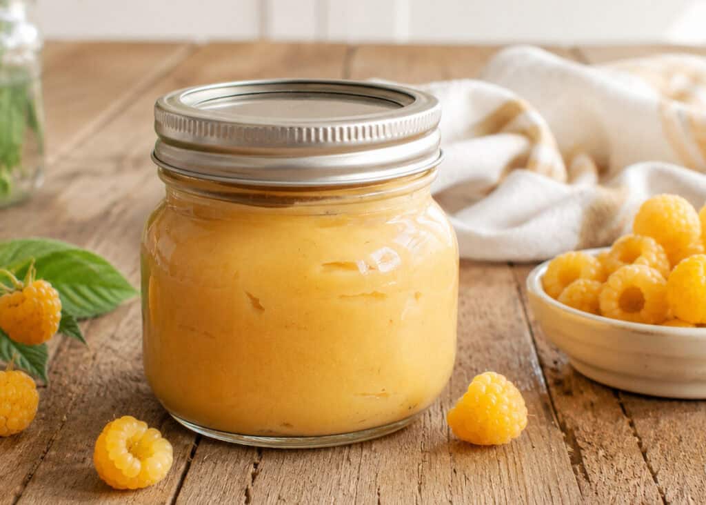 pint jar with a lid of golden raspberry curd. A small white bowl of golden raspberries is to the right of the jar, a white and golden striped kitchen towel is behind the jar, and 4 golden raspberries are scattered on the wooden table to the bottom and left of the jar.