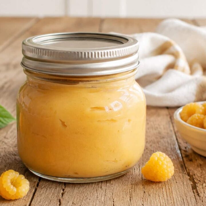 pint jar with a lid of golden raspberry curd. A small white bowl of golden raspberries is to the right of the jar, a white and golden striped kitchen towel is behind the jar, and 4 golden raspberries are scattered on the wooden table to the bottom and left of the jar.