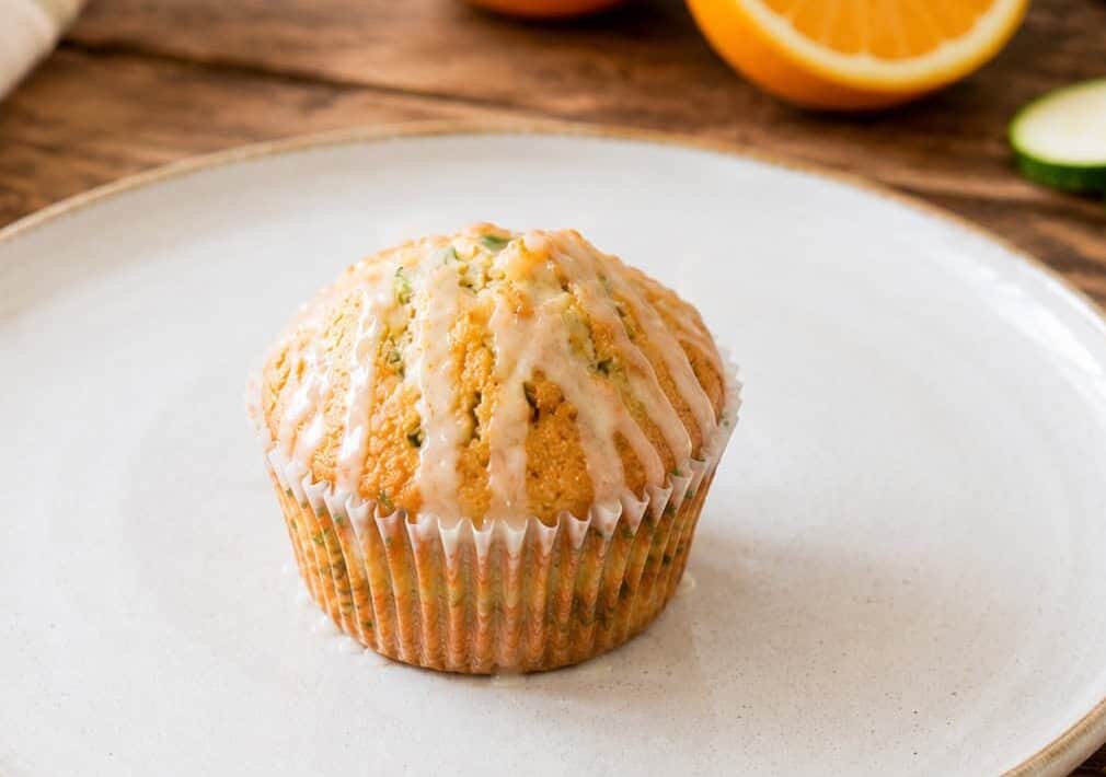 one orange cardamom zucchini muffin on a white plate sitting on a wooden table. A cut orange and sliced zucchini are on the table to the upper right.