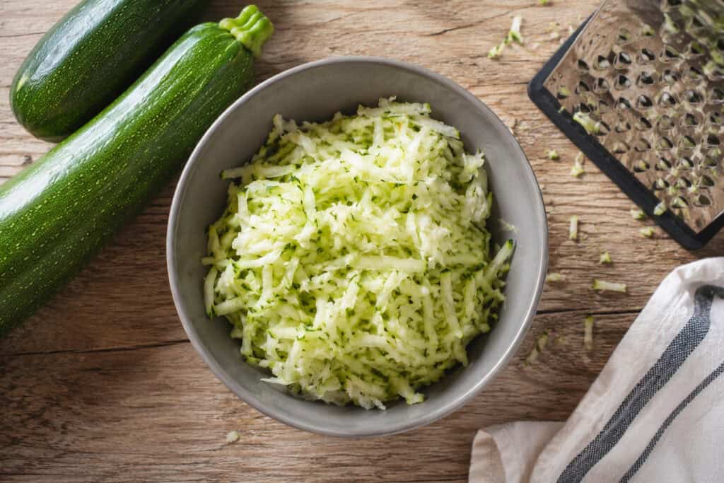 gray bowl of shredded zucchini on a wooden table with bits of shredded zucchini on it. To the upper left are two zucchini, to the upper right is a box grater with bits of zucchini still on it, and to the bottom right is a white and gray kitchen towel.