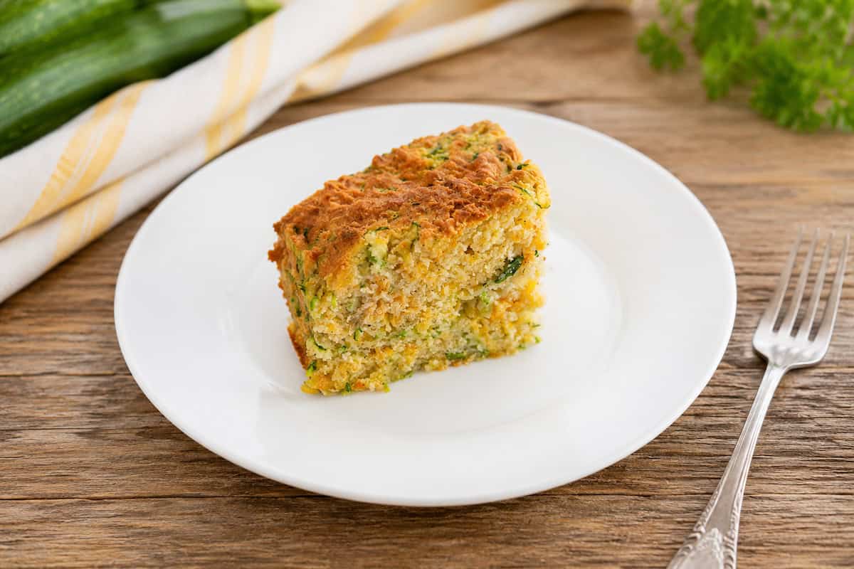 slice of zucchini cheddar breakfast squares on a white plate with a metal fork on a wooden table. 2 zucchini are stacked to the top left of the plate on a white and gold striped towel and a sprig of parsley is to the top right of the plate.