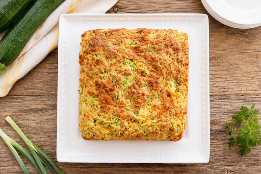 top down view of zucchini cheddar breakfast squares on a white plate sitting on a wooden table with 3 zucchini stacked on white and gold striped towel to the top left, green onions to the bottom left, and a stack of 4 white plates to the top right. A small sprig of parsley sitts to the bottom right.
