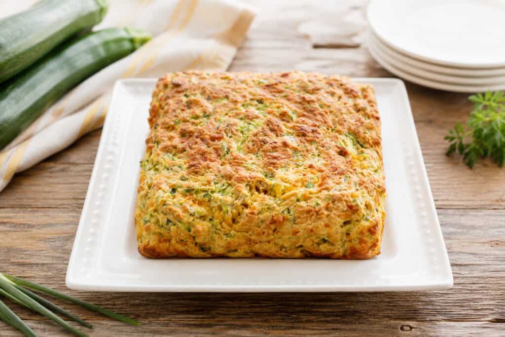 zucchini cheddar breakfast squares on a white plate sitting on a wooden table with 3 zucchini stacked on white and gold striped towel to the top left, green onions to the bottom left, and a stack of 4 white plates to the top right.