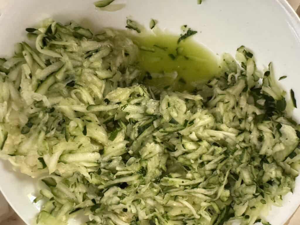 shredded zucchini in a white bowl that's been pushed over to show water pooled at the bottom of the bowl