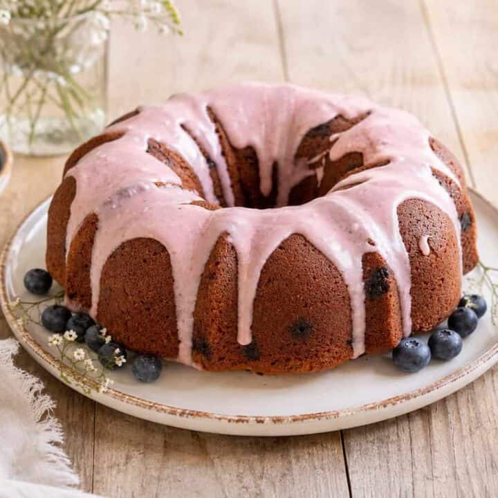 Blueberry zucchini bundt cake with blueberry glaze on a white plate sitting on a wooden table. There's a handful of blueberries on the plate with the cake and a small plate of blueberries to the left of the cake.