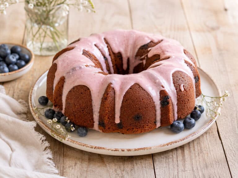 Blueberry zucchini bundt cake with blueberry glaze on a white plate sitting on a wooden table. There's a handful of blueberries on the plate with the cake and a small plate of blueberries to the left of the cake.