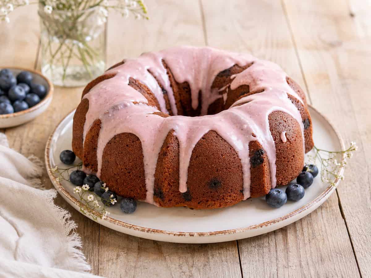 Blueberry zucchini bundt cake with blueberry glaze on a white plate sitting on a wooden table. There's a handful of blueberries on the plate with the cake and a small plate of blueberries to the left of the cake.