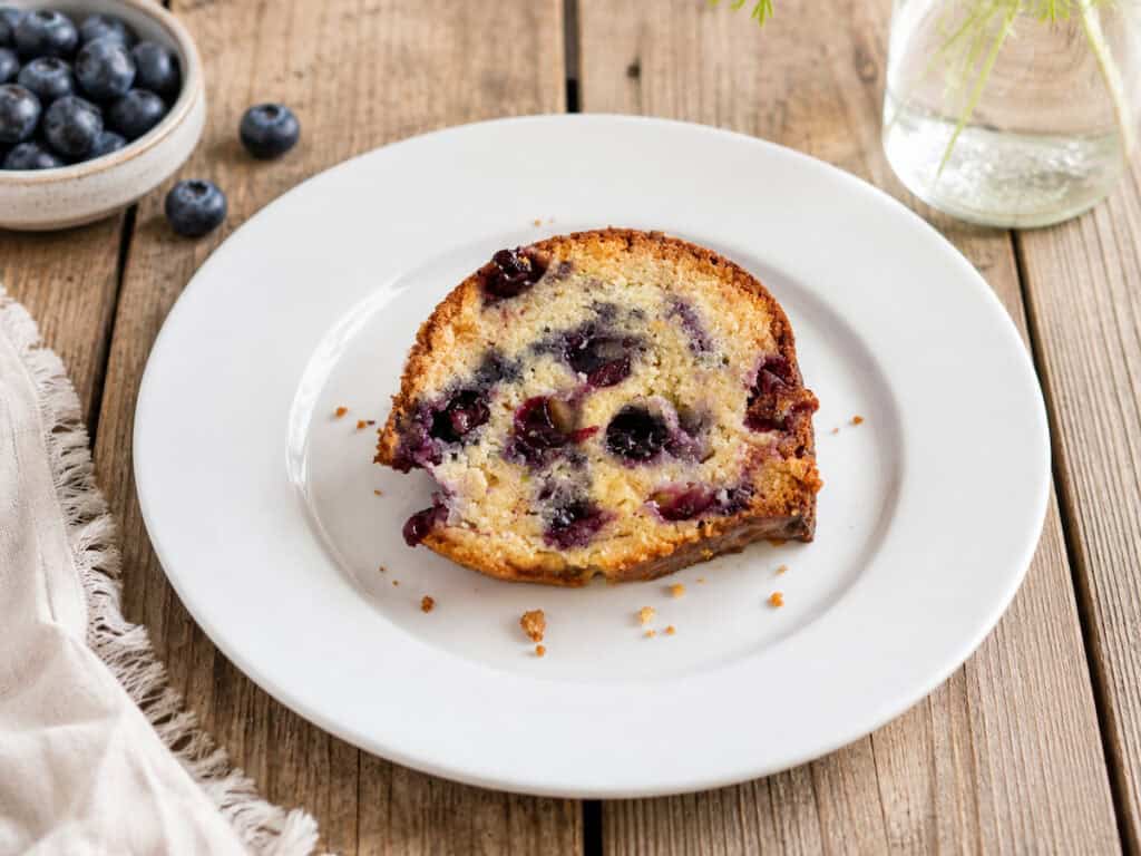 Blueberry zucchini bundt cake with blueberry glaze that has a slice taken out of it on a white plate sitting on a wooden table. There's a small plate of blueberries to the left of the cake.