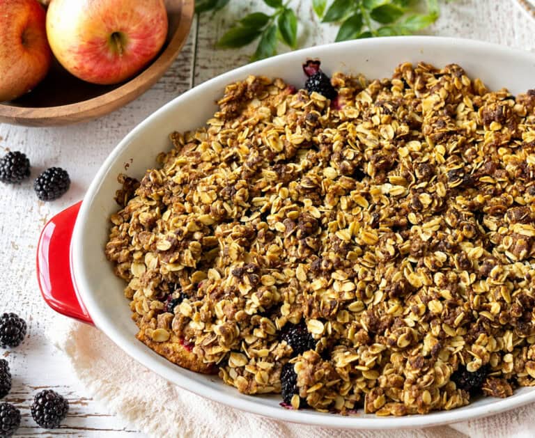 baked apple blackberry crisp in a white and red baking dish on a white table with a small wooden bowl of red apples in the upper left and a few blackberries scattered on the table to the middle and lower right