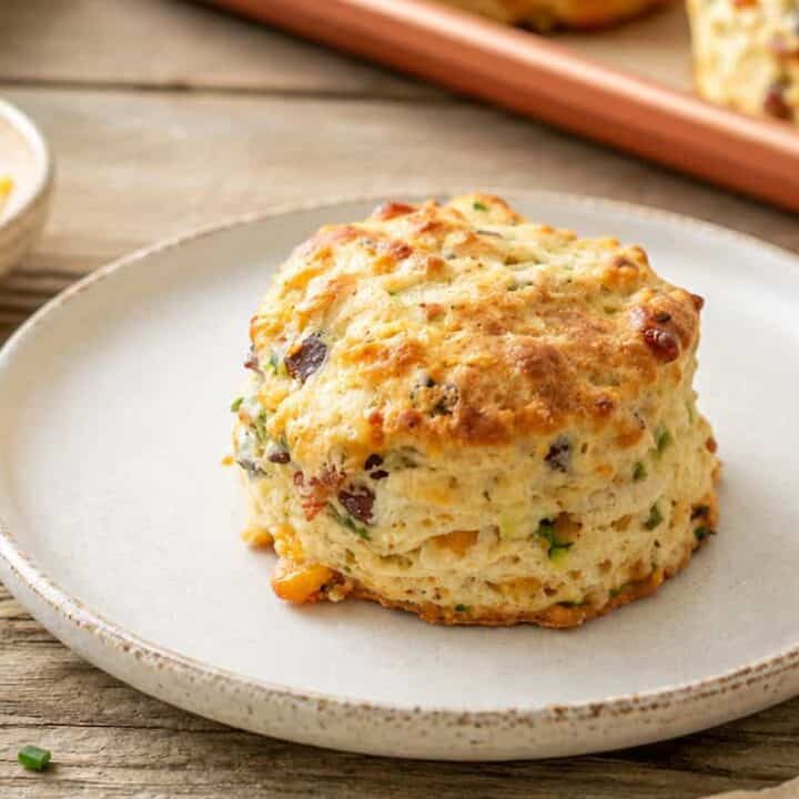 bacon cheddar zucchini biscuit on a white plate. A small white bowl of shredded cheese is to the upper left and the corner of the baking sheet with another biscuit on it is to the upper right