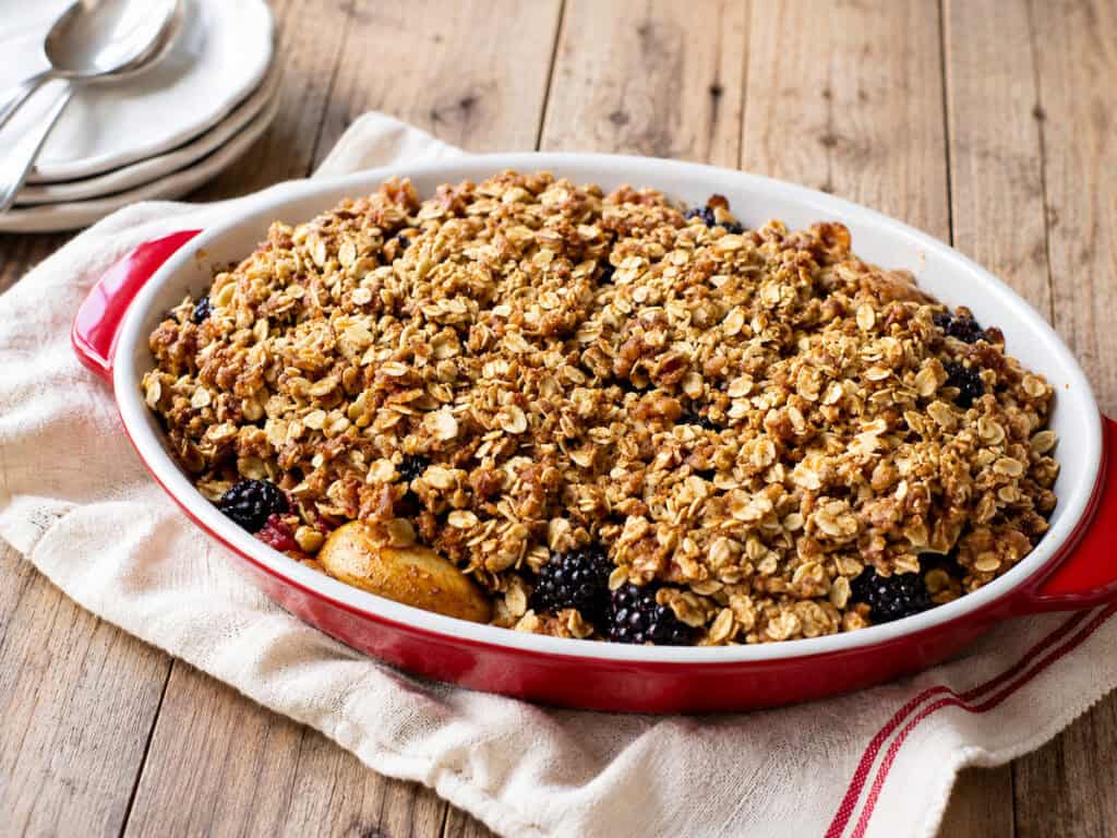 baked apple blackberry crisp in a white and red baking dish sitting on a white and red kitchen towel on a wooden table