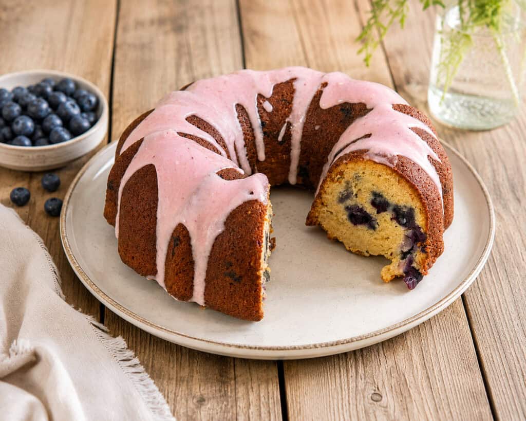 Blueberry zucchini bundt cake with blueberry glaze that has a slice taken out of it on a white plate sitting on a wooden table. There's a small plate of blueberries to the left of the cake.