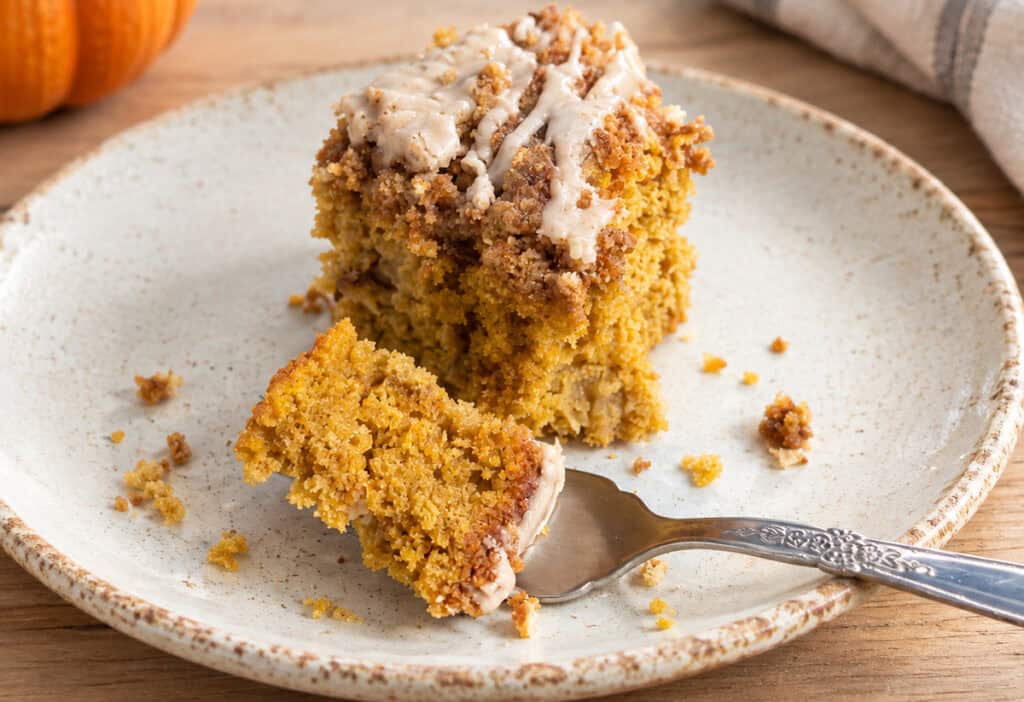Partially eaten slice of pumpkin apple crumb cake on a rustic gray white and brown plate. A fork is sitting on the plate in front of the slice with a bit of cake on the it. A sage green and white striped kitchen towel is to the upper right of the plate and a small portion of a pumpkin can be seen to the upper left of the plate.
