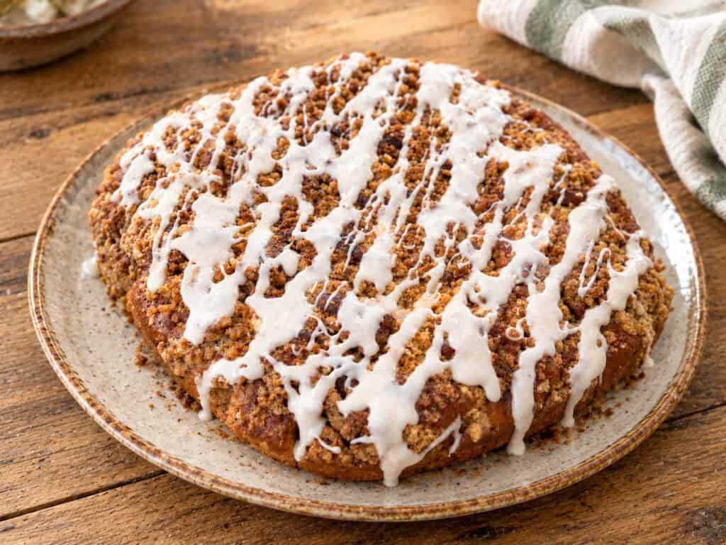 pumpkin apple crumb cake with cinnamon vanilla glaze on a rustic gray and brown plate sitting on a wooden table. A sage green and white striped linen cloth is to the upper right of the cake and a very small bit of a brown and gray bowl is to the upper left of the cake.