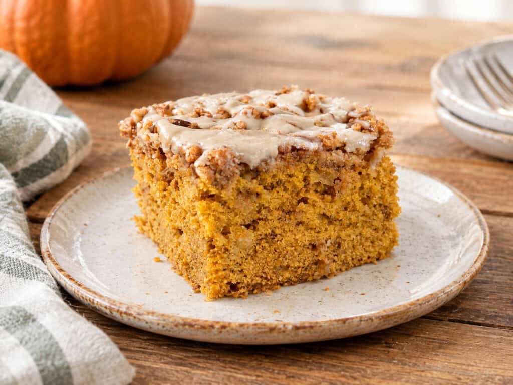 A slice of pumpkin apple crumb cake on a white and brown rustic plate, sitting on a wooden table. A sage green and white striped kitchen towel is to the lower left of the plate and you can see part of a pumpkin to the upper left. To the upper right are 2 more plates stacked on top of each other with a fork laying on the top plate.
