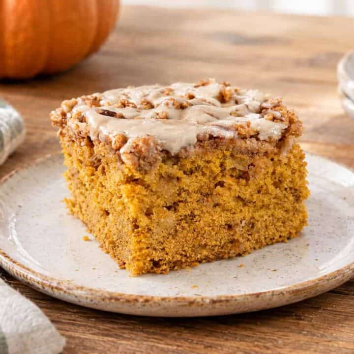 A slice of pumpkin apple crumb cake on a white and brown rustic plate, sitting on a wooden table. A sage green and white striped kitchen towel is to the lower left of the plate and you can see part of a pumpkin to the upper left. To the upper right are 2 more plates stacked on top of each other with a fork laying on the top plate.