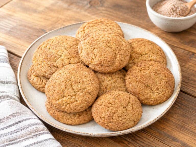 Yellow Squash Snickerdoodle Cookies on a white plate placed on a wooden table. A small bowl of cinnamon sugar mix is to the upper right and a white and silver striped kitchen towel is to the lower left.